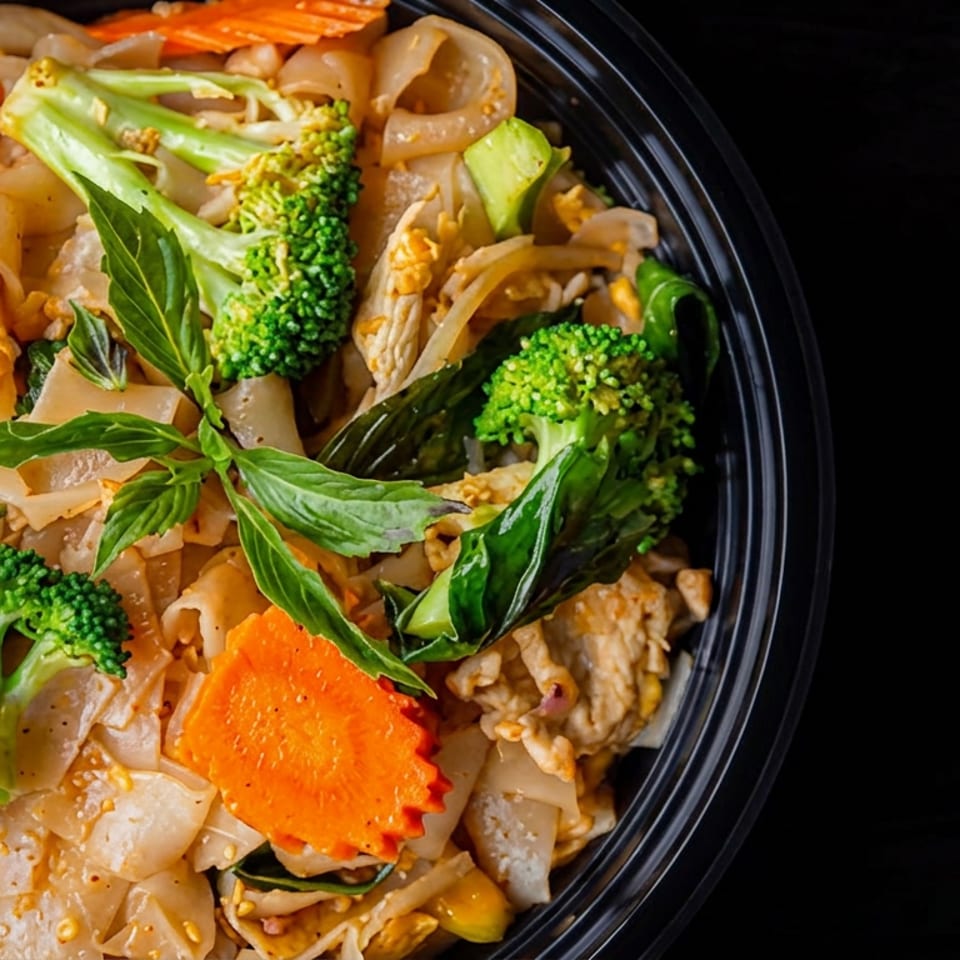 Bowl of stir-fried noodles with chicken, broccoli, carrots, onions, and leafy greens on a dark table.